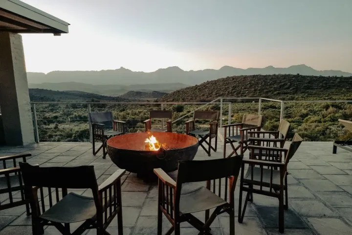 A steel fire bowl ringed by chairs on a stone terrace with hills beyond